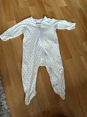 Vit pyjamas med gula hjärtan - Mysig vit pyjamas med långa ärmar och ben, täckt av små gula hjärtan. Dragkedja framtill som går snett ner mot benet för enkel påklädning. Tillverkad i mjukt bomullstyg och har inbyggda fötter för extra komfort.
