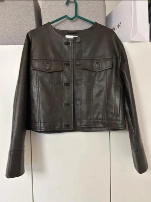 Brun skinnjacka från STOCKH LM Studio - Snygg brun skinnjacka från STOCKH LM Studio i storlek S. Jackan har rund halsringning, stora knappar framtill och två bröstfickor med lock. Rak, boxig passform och långa ärmar ger en modern look.