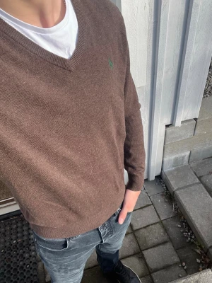 Brun stickad tröja från Polo Ralph Lauren - Snygg brun stickad tröja från Polo Ralph Lauren med v-ringning och diskret grön logga broderad på bröstet. Tröjan har långa ärmar och ribbade muddar vid ärmslut och nederkant. Perfekt att bära över en t-shirt för en avslappnad men stilren look.