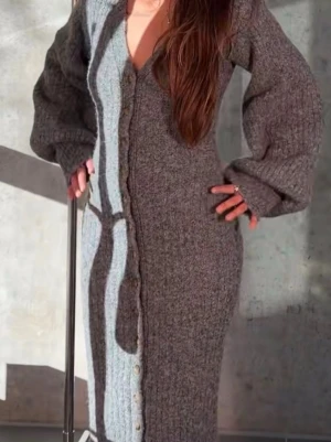 Grå stickad långklänning Adore - Mysig grå stickad långklänning från Adore med v-ringning och knappar längs hela framsidan. Klänningen har långa, lösa ärmar och är perfekt för kyligare dagar. Materialet är tjockt och mjukt stickat för extra komfort.