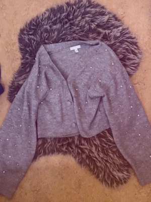 Grå kofta med pärldetaljer - Säljer en grå, croppad kofta med v-ringning och dekorativa pärlor över hela plagget. Koftan har långa, lite vidare ärmar och stängs med knappar framtill. Perfekt för dig som gillar en söt och trendig stil.