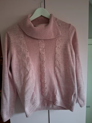 Ljusrosa polotröja med strukturmönster - Mysig ljusrosa stickad polotröja med bred ribbad krage och dekorativa strukturmönster framtill. Tröjan har långa ärmar och ribbade muddar vid ärmslut och nederkant. Perfekt för kalla dagar och enkel att matcha med jeans eller kjol.