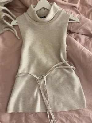 Beige ärmlös stickad poloväst - Säljer en beige stickad väst med polokrage och knytband i midjan. Västen är ärmlös och har en mjuk, slätstickad yta som ger en clean look. Perfekt att styla över en skjorta eller långärmad tröja för en trendig lager-på-lager stil.
