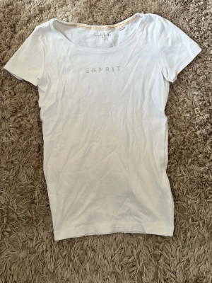 Vit t-shirt från Esprit med paljetter - Snygg vit t-shirt från Esprit med rund halsringning och korta ärmar. Framtill står det 'ESPRIT' i små paljetter som ger en extra touch. Tillverkad i mjuk bomull som känns skön mot huden. Perfekt till jeans eller kjol för en clean och fräsch look.