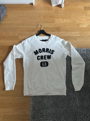 Vit sweatshirt Morris Crew 68 - Vit sweatshirt från Morris med texten 'Morris Crew 68' i mörkblått på bröstet. Tröjan har rund halsringning, långa ärmar och ribbade muddar vid ärmslut och nederkant. Tillverkad i mjukt bomullsmaterial för en bekväm känsla.