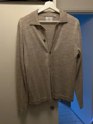 Beige cardigan från Bläck - Stilren beige cardigan från Bläck med klassisk krage och knäppning framtill. Tillverkad i ett tunt, mjukt material som passar perfekt för lager på lager. Långärmad modell med enkel design som funkar till många olika looks.