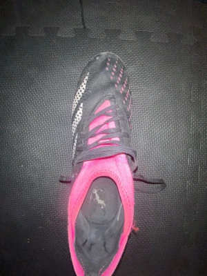 Adidas Predator svart/rosa fotbollsskor - Säljer ett par Adidas Predator fotbollsskor i svart och rosa med vita detaljer. Skorna har snörning och mönstrad ovandel för bättre bollkontroll. Syntetmaterial och dobbar för spel på gräs. 