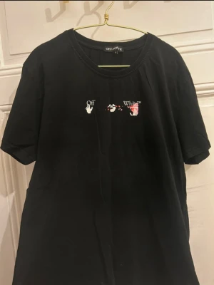 Svart Off-White t-shirt med tryck - Cool svart t-shirt från Off-White med unikt grafiskt tryck i vitt och rosa på ryggen och liten logga framtill. Klassisk rund hals och korta ärmar. Perfekt statement-plagg för dig som gillar streetwear. Scannar du QR Koden kommer du direkt till hemsidan! Säljer för 799 men kan sälja vid snabbköp!