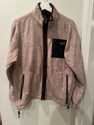 Patagonia fleecejacka pläd (burberry style) stl L herr - Patagonia jacka. Pläd (Burberry style) mikrofleece på utsidan, rutig flanellfoder i brunt och orange på insidan. Två looks i en. Bröstficka med dragkedja, två handfickor, dragsnöre i nederkant. Aldrig använd, utan lapp. Helt felfri.