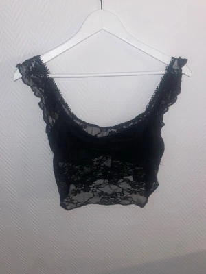 Svart spetsig croppad linnetopp - Säljer en svart croppad linnetopp i genomskinlig spets med breda axelband och volangkant. Perfekt till sommaren för en trendig look.
