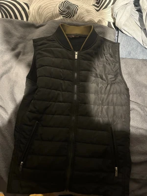 Svart dunväst med dragkedja - Snygg svart dunväst med quiltad design och hel dragkedja framtill. Två praktiska fickor med dragkedja på sidorna och kontrasterande krage i olivgrönt. Perfekt för lager-på-lager och streetwear-vibe. Dunvästen är från Zara 