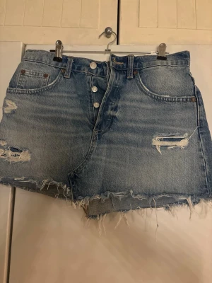 Blå jeansshorts med slitningar - Säljer ett par blå jeansshorts med råa kanter och slitna detaljer. Shortsen har hög midja, knappar framtill och klassiska fem fickor. Perfekta för sommaren och ger en avslappnad vibe.