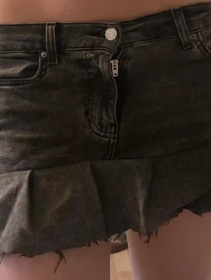 Svart jeanskjol  - Kort svart jeanskjol med rå, fransig kant och volangdetalj längst ner. Kjolen har klassiska jeansfickor fram och bak, bälteshällor och dragkedja med knapp framtill. Perfekt för en trendig och avslappnad look.