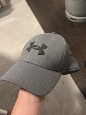 Grå keps från Under Armour - Snygg grå keps från Under Armour med böjd skärm och broderad svart logga framtill. Kepsen har klassisk passform och är tillverkad i ett slitstarkt tyg som känns sportigt och stilrent. Perfekt för dig som gillar enkel och clean stil.