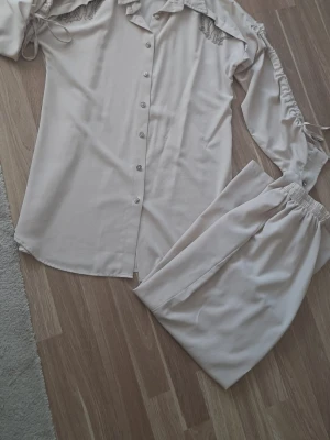 Beige set med spetsdetaljer - Säljer ett beige set med långärmad body och matchande byxa. Bodyn har knappar framtill, spetsdetaljer vid axlarna och snörning på ärmarna. Materialet känns mjukt och lätt, perfekt för en stilren look.