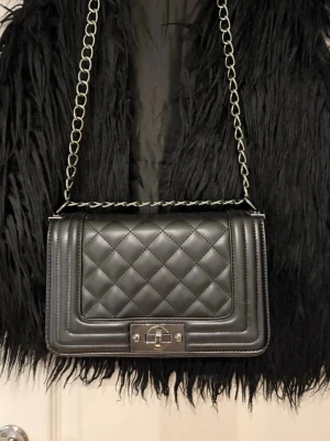 Svart quiltad axelväska med kedja - Snygg svart axelväska i quiltat mönster med silverfärgad kedjerem och vridlås. Väskan har en stilren, rektangulär form och är tillverkad i konstläder. Perfekt för dig som gillar en klassisk men trendig look.