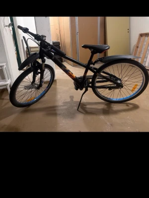 Svart mountainbike med stänkskärmar - Säljer en svart mountainbike med orange detaljer och blå reflexer på hjulen. Cykeln har stänkskärmar fram och bak, samt stöd. Sadel och handtag är svarta och ramen har en sportig design. Perfekt för dig som vill ha en cool cykel till vardagsäventyr.