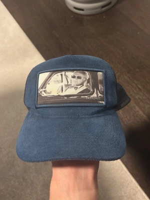 Blå truckerkeps med motiv från Defadati - Snygg blå truckerkeps från Depadati med ett svartvitt motiv framtill som föreställer två personer i en bil. Kepsen har mesh på sidorna och justerbart spänne bak i läderimitation. Perfekt för dig som vill sticka ut med en unik accessoar.