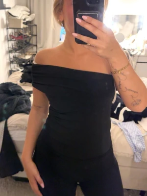 Svart offshoulder topp - Snygg svart offshoulder topp med tight passform och draperad design. Toppen är axelbandslös och har en stilren look som passar perfekt till många olika outfits. Materialet känns mjukt och stretchigt, vilket gör den bekväm att bära.