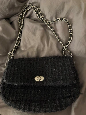 Svart axelväska med kedjerem - Snygg svart axelväska i tweed-liknande tyg med silverfärgad kedjerem och vridlås. Väskan har en klassisk, rundad form och lock. Perfekt för att lyfta din outfit med en trendig detalj.