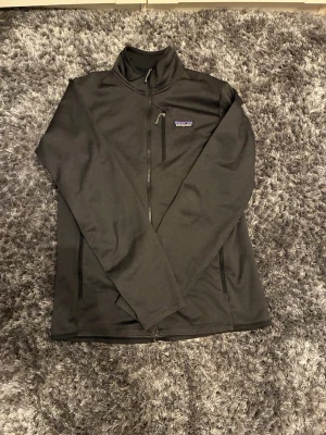Mörk grå ziptröja från Patagonia - Snygg mörk grå ziptröja från Patagonia med hög krage och dragkedja framtill. Tröjan har tre fickor med dragkedja och en liten logga på bröstet. Tillverkad i ett mjukt och stretchigt material.