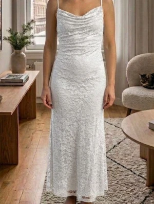 Vit lång spetsklänning med smala band - Elegant långklänning i vit spets med smala axelband och lätt draperad byst. Klänningen har figurnära passform och faller mjukt längs kroppen. Perfekt för sommarens speciella tillfällen.