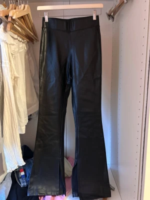 Svarta utsvängda skinbyxor - Säljer ett par svarta skinbyxor med utsvängda ben och hög midja. Byxorna har slits framtill vid bensluten och en clean look utan synliga fickor eller detaljer. 