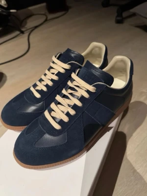 Marinblå sneakers med mockadetaljer - Snygga marinblå sneakers med ovandel i både mocka och läder, beige snören och vit insida. Skorna har en klassisk låg siluett och gummisula i brunt. Perfekta för dig som gillar stilrena och tidlösa sneakers.