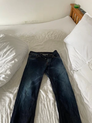 Mörkblå jeans från Nudie Jeans - Säljer ett par mörkblå jeans från Nudie Jeans med klassisk femficksmodell och raka ben. Jeansen har kontrastsömmar och Nudie-logga på bakfickan. Tillverkade i slitstark denim, perfekta för en avslappnad och snygg stil. W31 L32