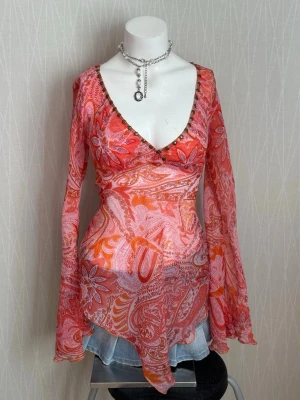 Fairy Mönstrad omlottopp med vida ärmar - Trendig omlottopp i tunnt, transparent material med paisleyinspirerat mönster i rosa, orange och rött. Toppen har v-ringning med små nitar, långa utsvängda ärmar och asymmetrisk nederkant. Perfekt för dig som gillar boho-stil.