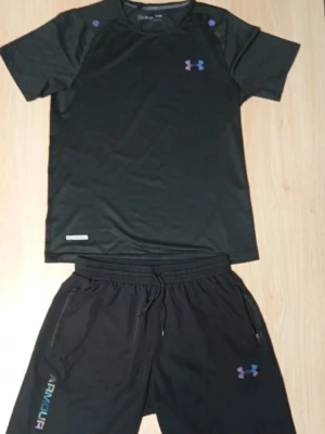Under Armour svart träningsset - Snyggt svart träningsset från Under Armour med matchande t-shirt och shorts. Setet har diskreta loggor i lila och blått, samt reflekterande detaljer på både tröja och byxa. Tillverkat i ett lätt och snabbtorkande funktionsmaterial som passar perfekt till träning.