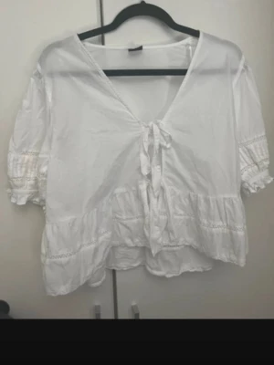 Vit blus med knyt och volanger från Gina trico - Söt vit blus med v-ringning och knyt framtill. Blusen har volangdetaljer och trekvartsärmar med resår och spetsinslag. Perfekt för en avslappnad och trendig look. Tillverkad i ett lätt bomullsliknande material. Från Gina trico. Köpt för 450