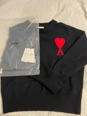 Svart stickad tröja från Ami Paris - Säljer en svart stickad tröja från Ami Paris med röd hjärtlogga och bokstaven A på bröstet. Tröjan har rund halsringning, långa ärmar och breda ribbade muddar vid ärmslut och nederkant. Perfekt för dig som gillar stilrena plagg med en twist.