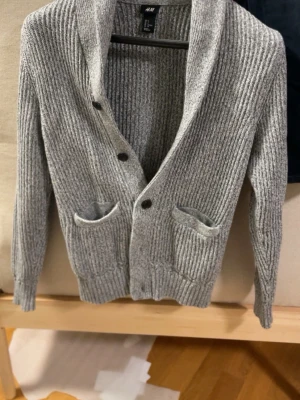 Grå ribbstickad kofta från H&M - Snygg grå ribbstickad kofta från H&M med sjalkrage och tre knappar framtill. Koftan har två framfickor och ribbade muddar vid ärmslut och nederkant. Perfekt för lager på lager och en chill look.
