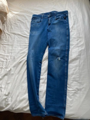 Blå jeans Replay Anbass herr 31 - Säljer ett par blå jeans från Replay, modell Anbass. De har en klassisk femficksdesign, raka ben och slitningar på låret för en trendig look. Jeansen är i stretchigt material för extra komfort och har snygga detaljer vid fickorna. Ett litet hål på vänstra knät som man set på bild 5 och 6. 