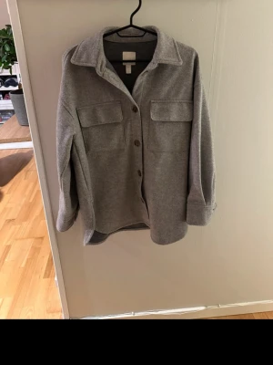 Grå kappa med fickor - Stilren grå overshirt med krage och knäppning framtill. Två stora bröstfickor med lock ger en trendig look. Overshirten är i ett mjukt, filtliknande material och har långa ärmar med breda muddar. Perfekt att bära lager på lager för en avslappnad stil.