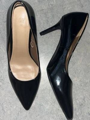 Svarta blanka klackskor - Säljer ett par klassiska svarta pumps i storlek 38. Skorna har spetsig tå, smal klack på ca 6-8cm och har ett glansigt lackmaterial. Splitternya, endast provat inomhus men har ej kvar förpackningen. Kan självklart diskutera pris🩷