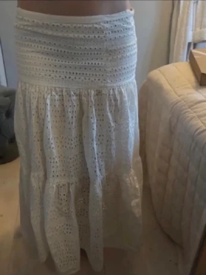 Vit långkjol i broderad bomull - Säljer en vit långkjol i broderad bomull med vackra spetsdetaljer och hålmönster. Kjolen har en bred, smockad midja och faller i flera volanger för en luftig och romantisk look. Perfekt för sommare!
