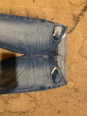 Blå bootcut jeans från Diesel - Säljer ett par klassiska blå bootcut jeans från Diesel med slitningar och tvättade detaljer. Jeansen har fem fickor, dragkedja och knapp framtill samt en liten logga på fickan. Perfekta för en avslappnad och trendig look.