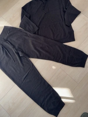Mörkgrått loungewear-set med hoodie - Mysigt loungewear-set i mjukt material, består av en mörkgrå hoodie med huva och dragsko samt matchande byxor med resårmidja och dragsko. Perfekt för chill dagar hemma. Setet har en avslappnad passform och långa ärmar och ben. ÄKTA KASHMIR OCH ULL!