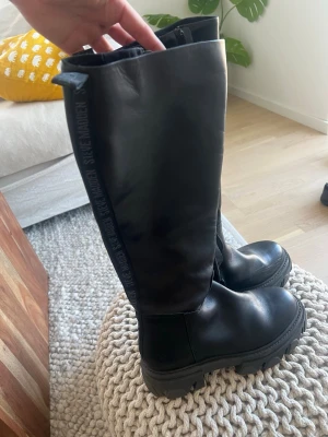 Svarta höga boots från Steve Madden - Säljer ett par svarta höga boots från Steve Madden med chunky sula och rund tå. Stövlarna är i slätt skinnliknande material och har diskret logga längs kanten. Perfekta för en edgy och trendig look.