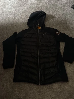 Svart pufferjacka med huva - Snygg svart paraymperjacka med huva och dragkedja framtill. Jackan har quiltade partier på bröst och rygg, samt en bröstficka med dragkedja. Ärmar och sidor är i stretchigt material för extra komfort. Märke syns på ärmen. Storlek på jackan är 176 cm