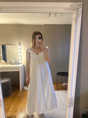 Vit långklänning med volangdetaljer - Vit snygg långklänning med volangkant vid axlar och urringning. Klänningen har en lös passform och djup rygg, vilket ger en luftig och elegant känsla. Perfekt för sommarens alla tillfällen.