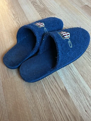 Mörkblåa tofflor med brodyr - Mysiga mörkblåa tofflor i mjukt tygmaterial med broderad detalj framtill. Öppen häl och klassisk slip-in modell, perfekta att glida i hemma. Brodyren har ett märke med text och rödvita detaljer.