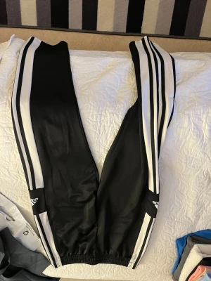 Svarta Adidas mjukisbyxor med vita ränder - Säljer ett par klassiska svarta Adidas mjukisbyxor med de ikoniska tre vita ränderna längs benen. Byxorna har resår i midjan och vid bensluten samt Adidas-logga på båda benen. Perfekta för en sportig och avslappnad stil.