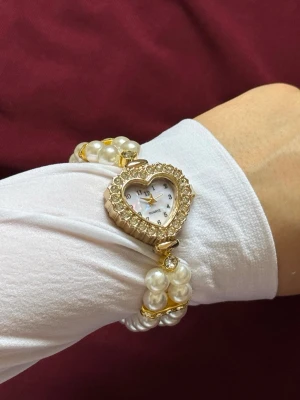 Pärlklocka med hjärtformat ur - Unik klocka med hjärtformat ur i guldton och glittrande stenar runt boetten. Armbandet består av dubbla rader vita pärlor och guldfärgade detaljer. Perfekt accessoar för dig som gillar romantisk och elegant stil.