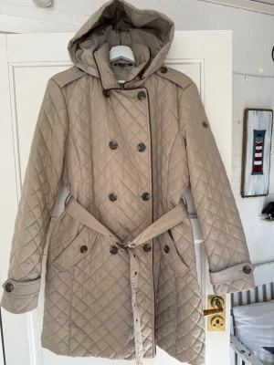 RALPH LAUREN quiltad trenchcoat med huva - Ralph Lauren  beige trenchcoat med quiltat mönster och avtagbar huva. Jackan har dubbelknäppning, bälte i midjan och dekorativa knappar vid ärmsluten. Perfekt för dig som vill ha en klassisk men modern look. Passar till både vardag och cityhäng.