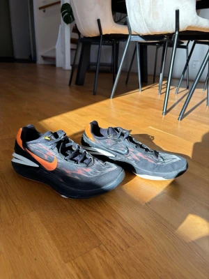 Nike svarta sneakers  - Snygga svarta Nike sneakers med orange swoosh och detaljer i grått och vitt. Ovandelen är i mesh för bra andningsförmåga och sulan är tjock och dämpad för extra komfort. Skorna har lågt skaft och svarta skosnören. Perfekta för en sportig och trendig look.