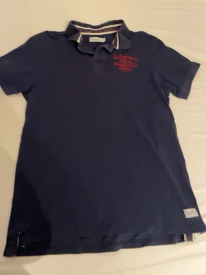 Mörkblå pikétröja med röd brodyr - Snygg mörkblå pikétröja med krage och korta ärmar. Tröjan har röd brodyr på bröstet och diskreta detaljer vid kragen. Tillverkad i mjuk bomull som känns skön mot huden. Perfekt för en avslappnad men ändå stilren look.