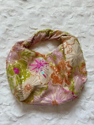 Blommig handväska med paljetter - Unik handväska i beige tyg med broderade blommor i grönt, orange, rosa och lila. Väskan är dekorerad med paljetter och pärlor som ger extra glans. Rundad form med vadderat handtag och dragkedja upptill. Perfekt för dig som gillar färg och detaljer.